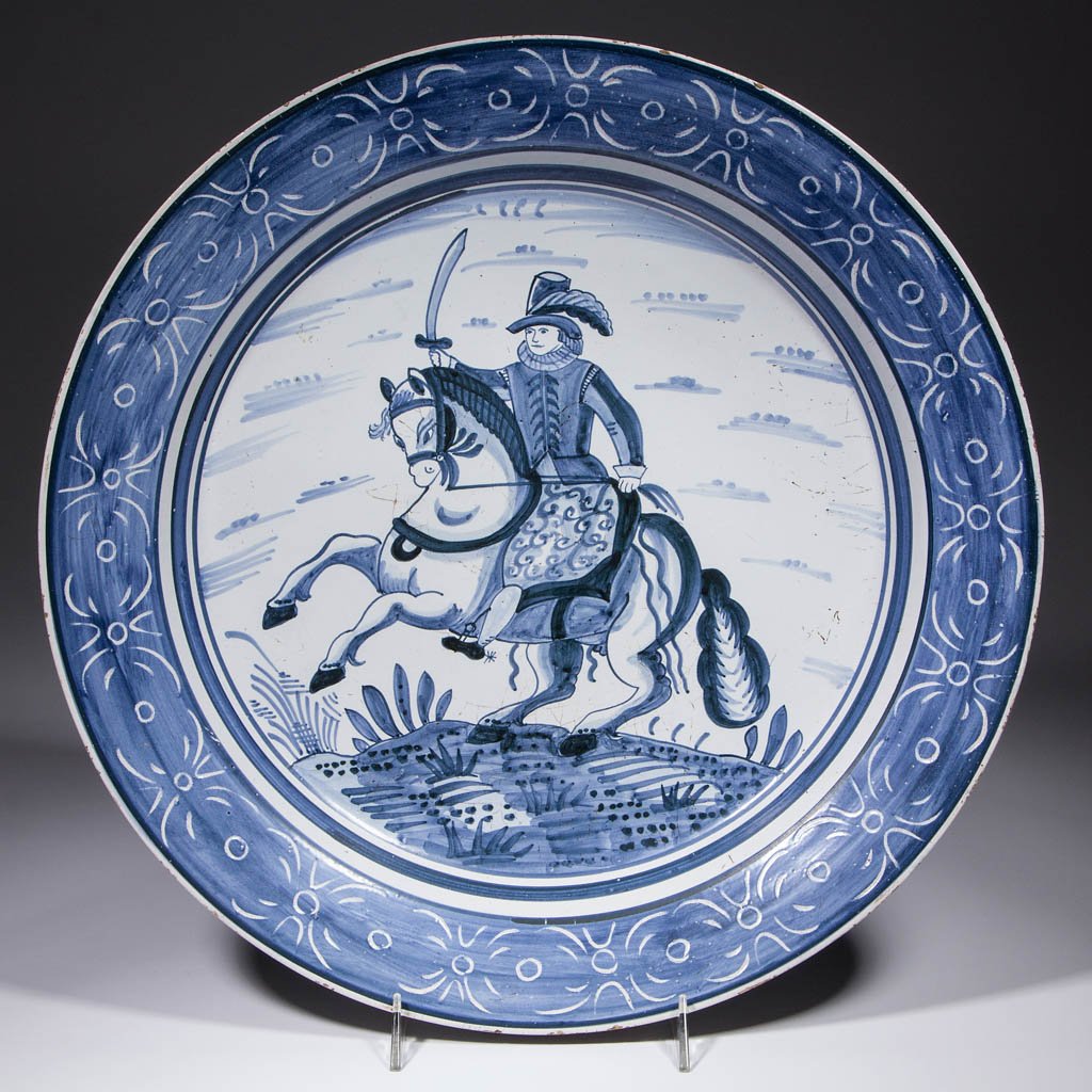 DUTCH DELFT TIN-GLAZED EARTHENWARE CHARGER: DUTCH DELFT TIN-GLAZED EARTHENWARE CHARGER, having a central image of a man on a horse in elaborate clothing with a sword, sgraffito decoration on rim, pierced foot rim with metal hanger. Intertwined