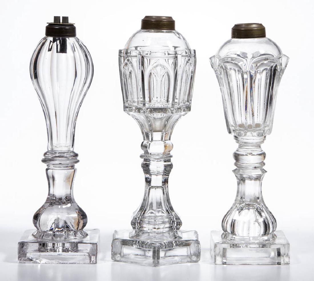 ASSORTED PRESSED WHALE OIL / FLUID STAND LAMPS, LOT OF: ASSORTED PRESSED WHALE OIL / FLUID STAND LAMPS, LOT OF THREE, colorless, comprising a Loop/Leaf, a cut flutes example, and a Gothic Arch example; each with a period brass collar. One example fitted wi