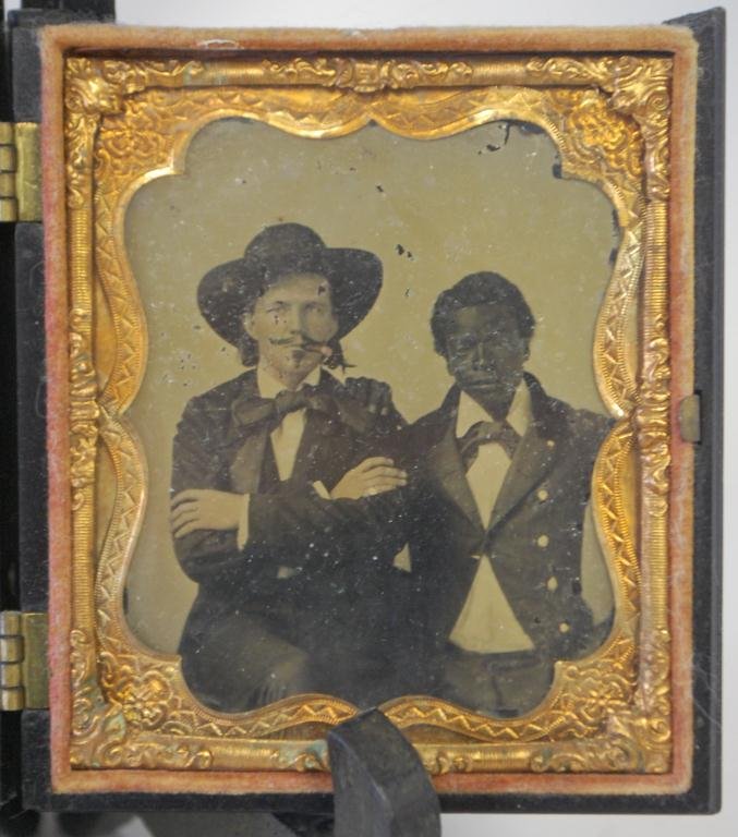 Civil War Era Ambrotype Black and White Men: Ambrotype of a white man in broad brimmed hat and suit, with cigar and crossed arms, flanked by a younger black man, also in suit, with his hand on his companion's shoulder. In embossed case, some dam