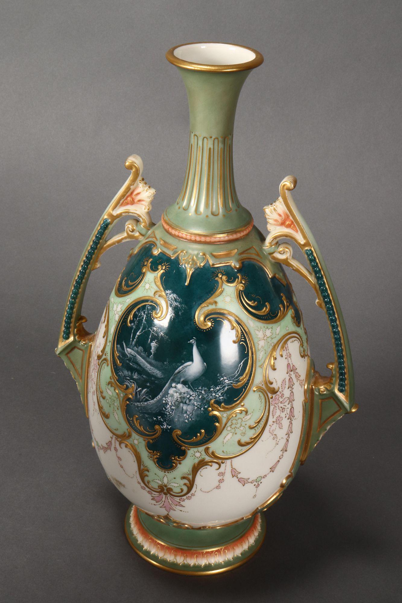 Royal Worcester Twin Handled Porcelain Vase,: of bulbous form with a slender tapering neck, decorated with gilt c-scroll cartouche displaying two peacocks in a green landscape, surrounded by floral motifs on a pale green and cream ground, accente