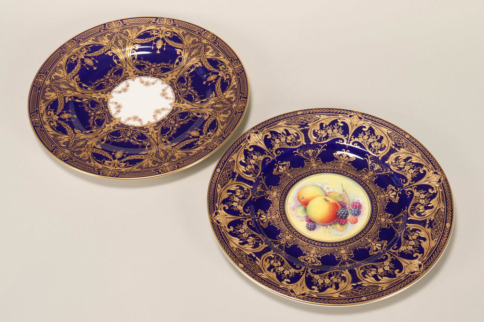 Two Royal Worcester Cabinet Plates,: one with central hand painted roundel depicting fruits and berries, signed P.Platt, set within cobalt and gilt decorated borders, diameter 27cm, the other with central white reserve within cobalt and 
