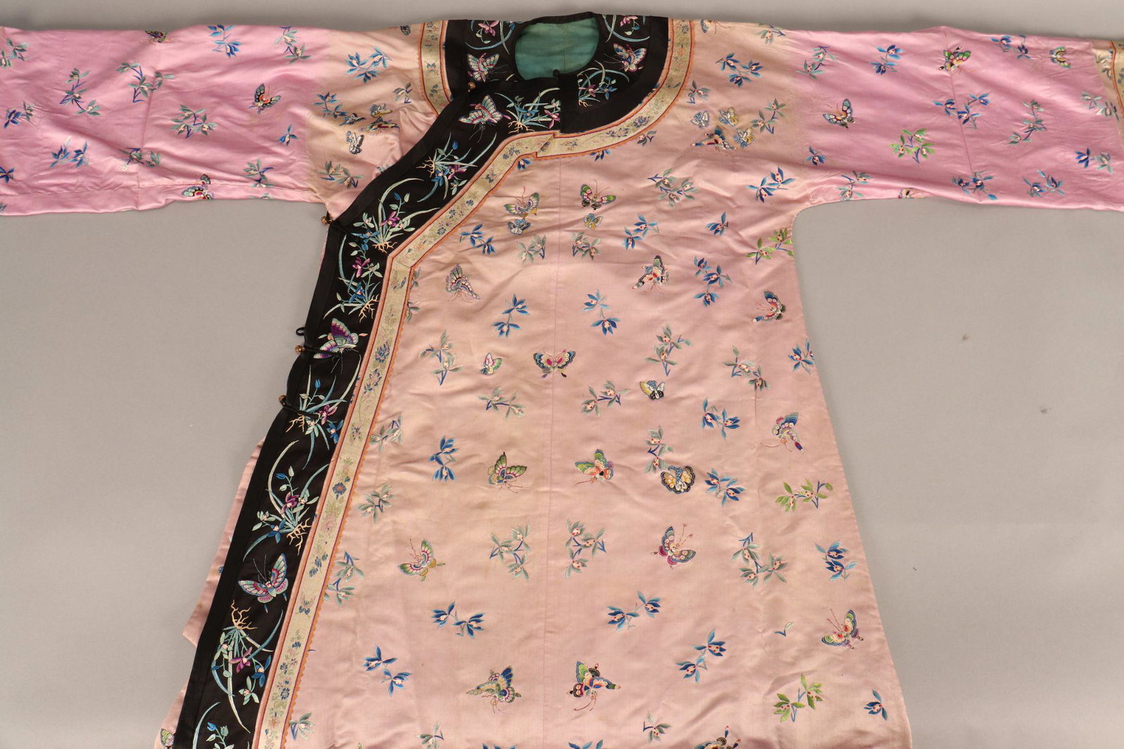 Chinese Pink Silk Manchu Lady's Informal Robe,: Changyi, Late Qing Dynasty, embroidered in satin stitch and brightly coloured butterflies hovering over scattered orchid sprays, enclosed by a black silk border similarly worked with pairs of orchid p