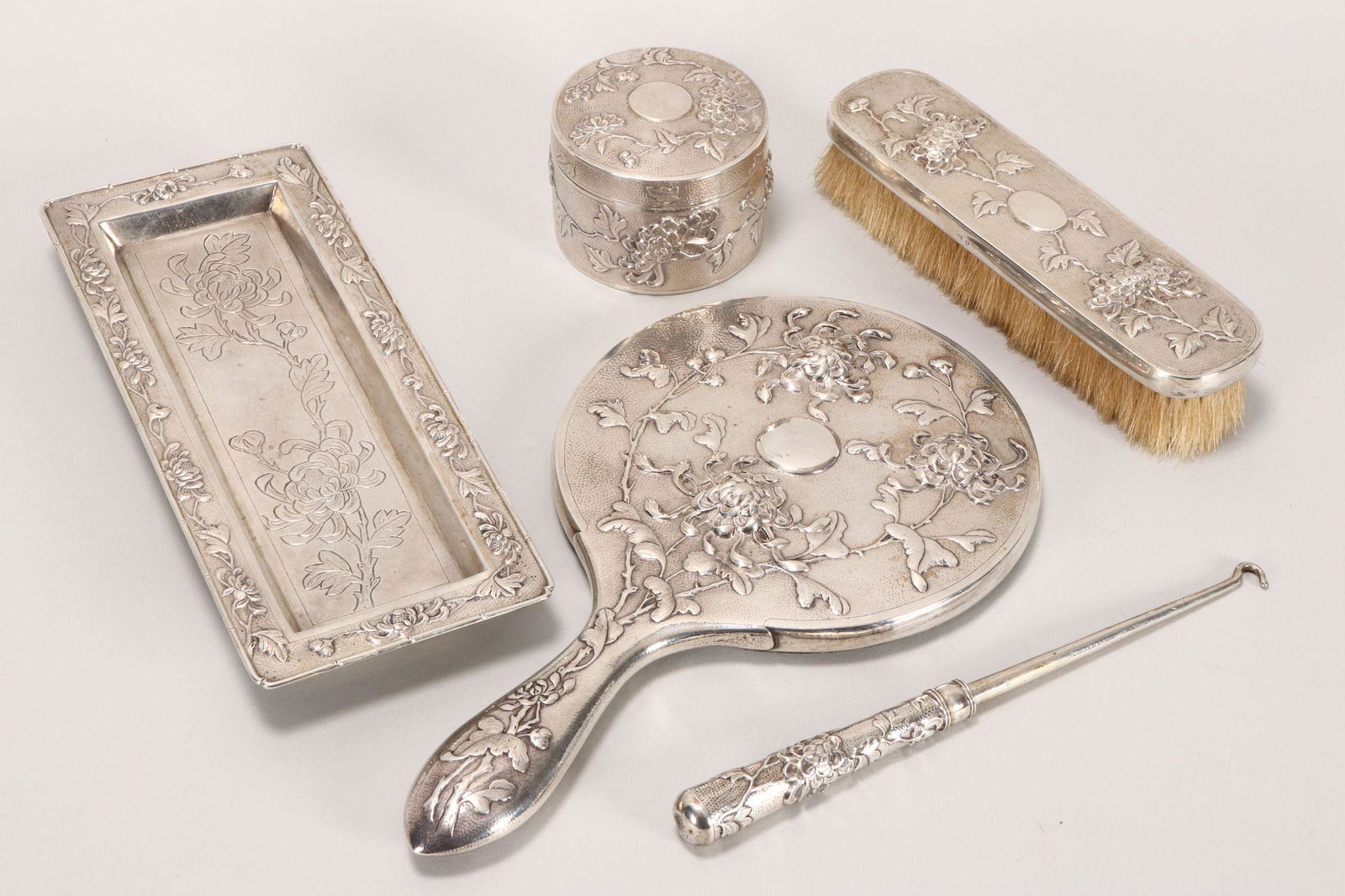 Chinese Export Silver Five Piece Dressing Table: Set, all marked HC for Hung Chong, comprising hand mirror, clothes brush, box and cover, rectangular footed tray and button hook, all decorated with peonies and foliage in relief (5)