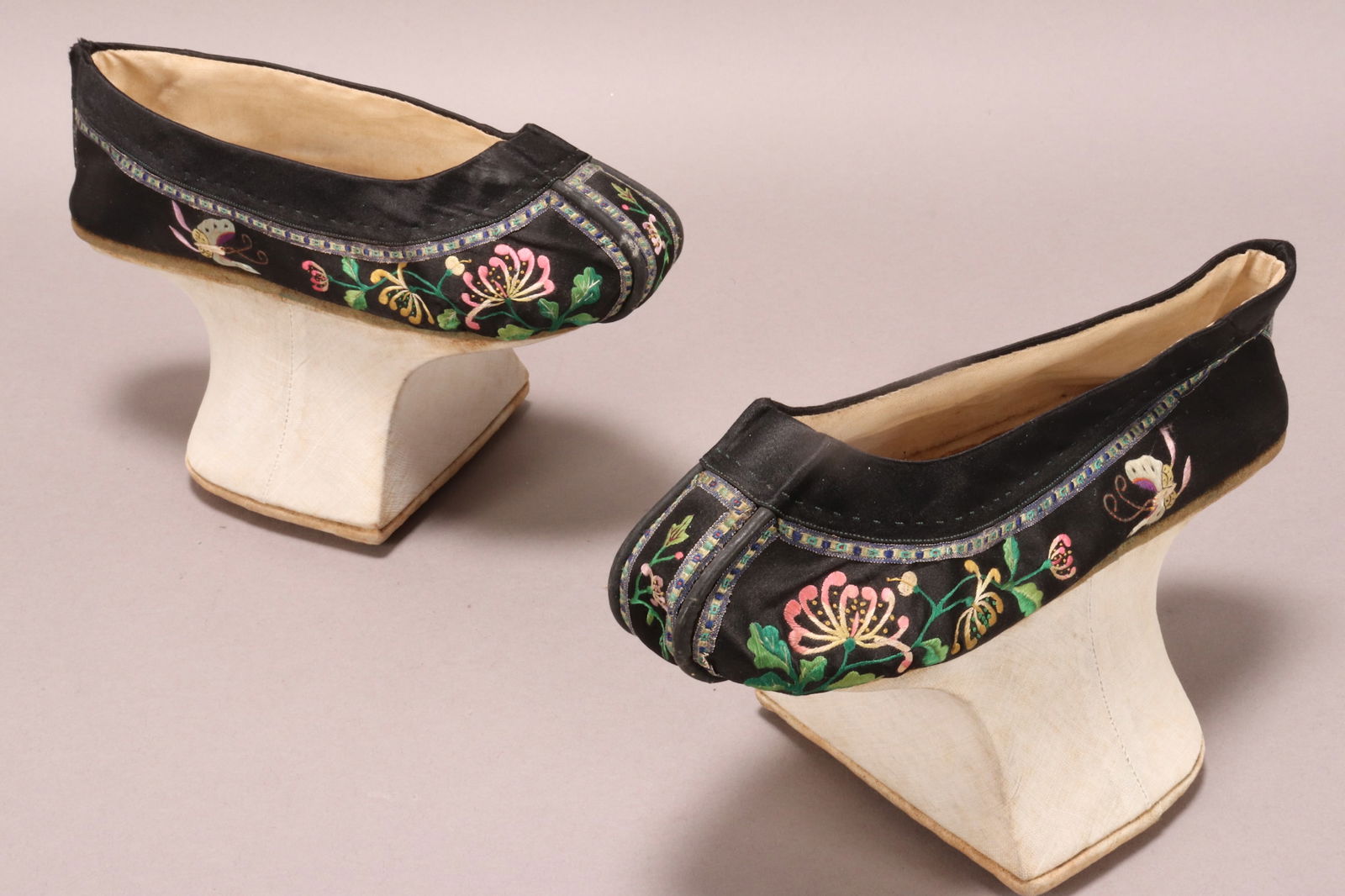 Pair of Chinese Qing Dynasty Manchu Horse-Hoof Shoes,: in black silk, decorated with embroidered chrysanthemums and butterflies with couched antennae, with a ribbon border, shoe heel is constructed from wood with a fabric covering and padded sole, in tone