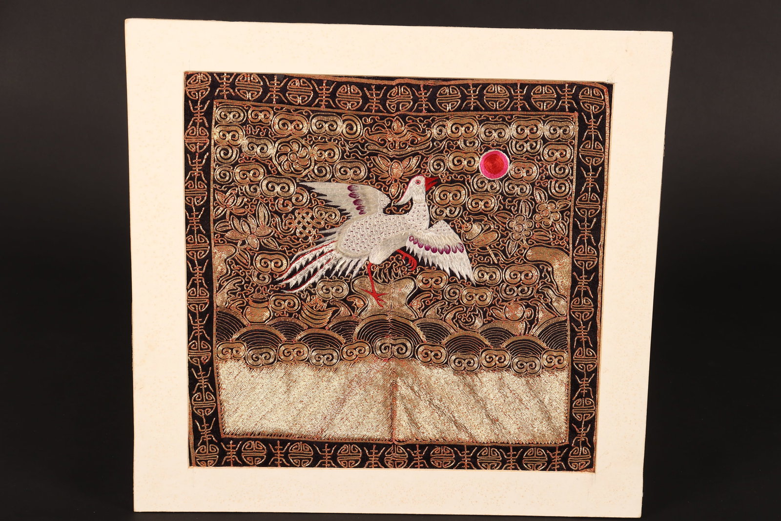 Chinese Embroidered Rank Badge,: of square form, mounted on card, depicting a central crane standing on one foot on a raised platform, with a surrounding scene containing the sun to upper right, scrolling motifs, flowers, foliage and