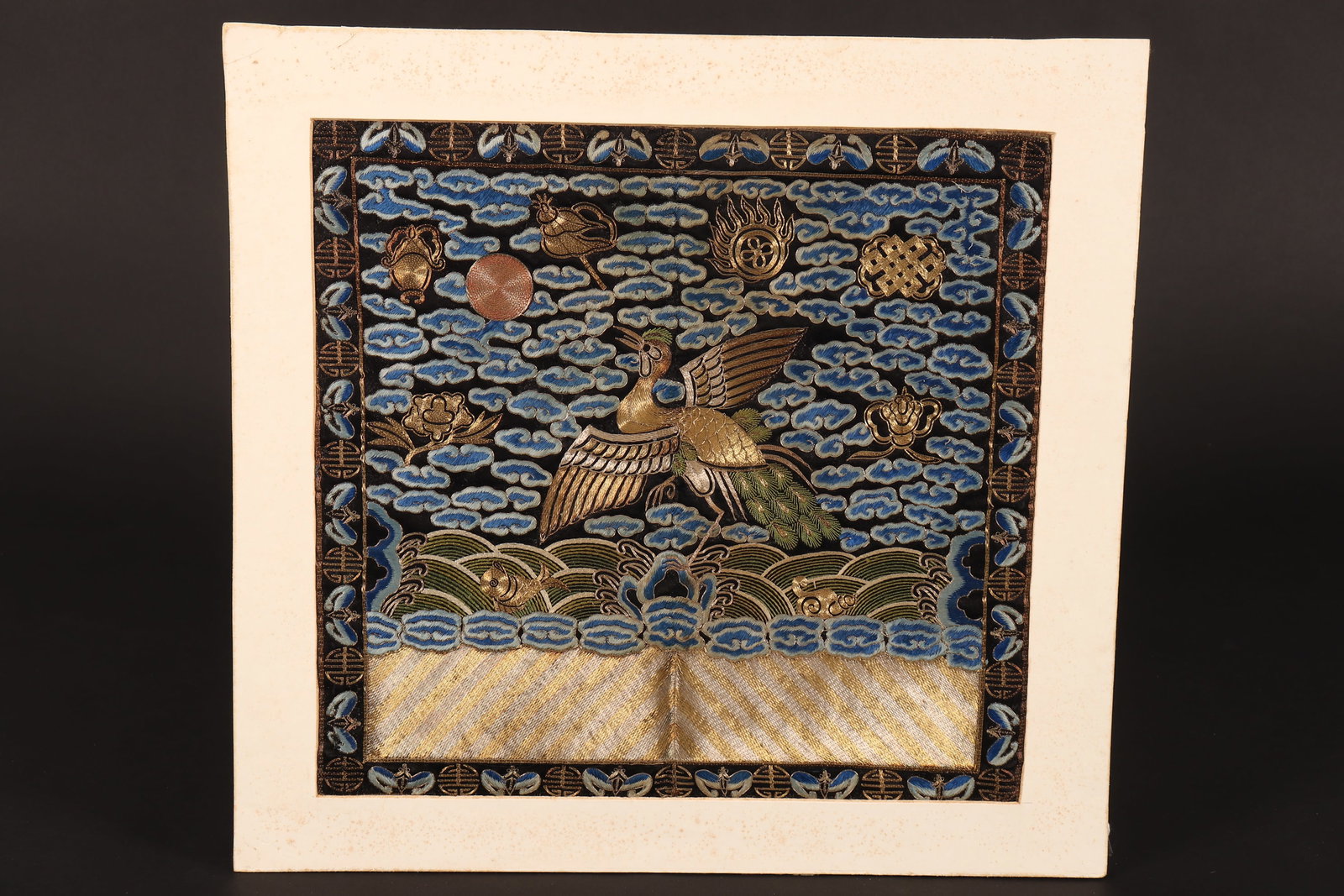 Chinese Embroidered Rank Badge,: of square form, mounted on card, depicting a central crane standing on one leg on a raised platform, with a surrounding scene containing the sun to upper right, stylised clouds, flaming pearl, waves, 