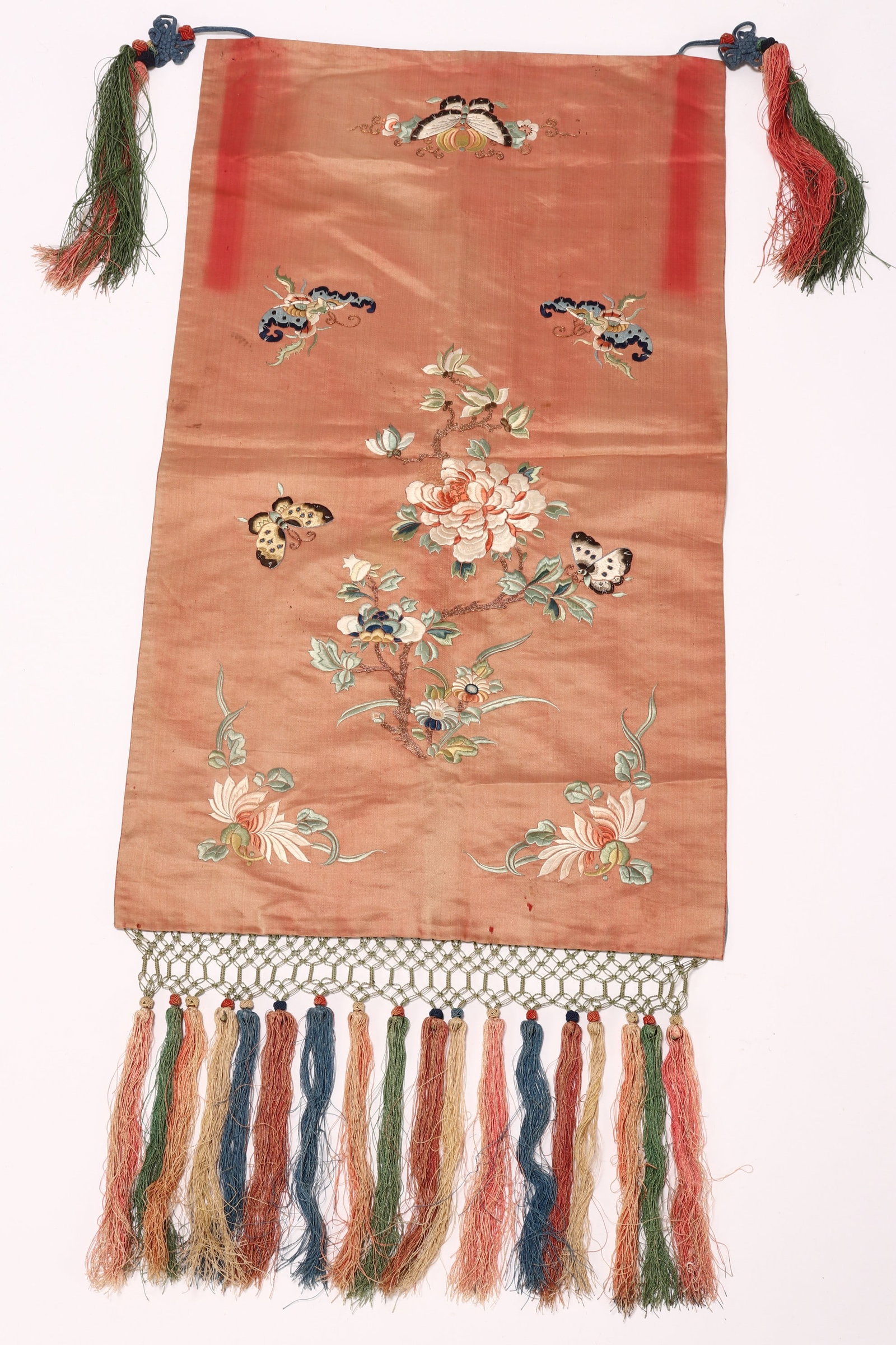 Chinese Peach Satin Embroidered Textile,: embroidered with peonies, butterflies and foliage, in tones of blue, green and brown, with tassels to each end, 64cm x 37cm (not including tassels)