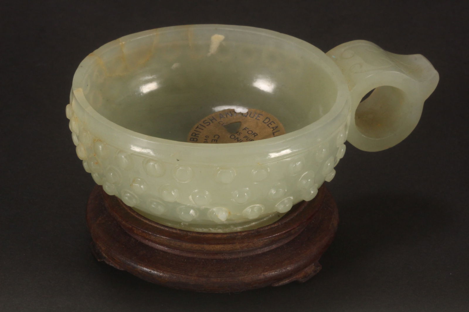 Chinese Qing Dynasty Pale Jade Cup,: of circular form, with dimpled exterior and looped handle, raised on wooden stand, with The British Antique Dealers Association label to interior of bowl, height 3cm, diameter 6cm, length 8.5cm (inclu