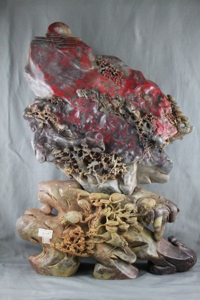 Well Carved Chinese Chicken Blood Stone Boulder,: finely carved on both sides with mountain landscape of storks amongst trees, in tones of red, grey and black, resting upon a base carved with fern tree motifs, in tones of yellow, h 39cm. Provenance: 