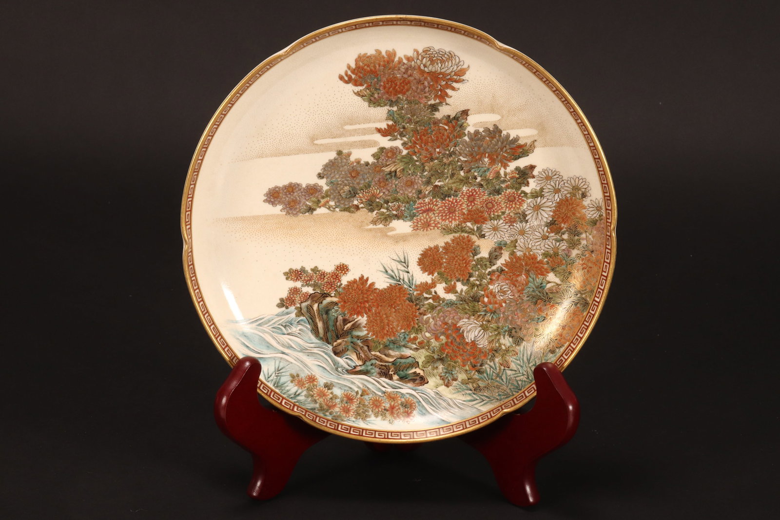 Fine Japanese Meiji Period Satsuma Plate,: by Kizan, of circular form with pinched rim, finely decorated with a central scene of flowering trees next to a rocky formation and flowing stream, encased by a Greek key border, in tones of green, bl