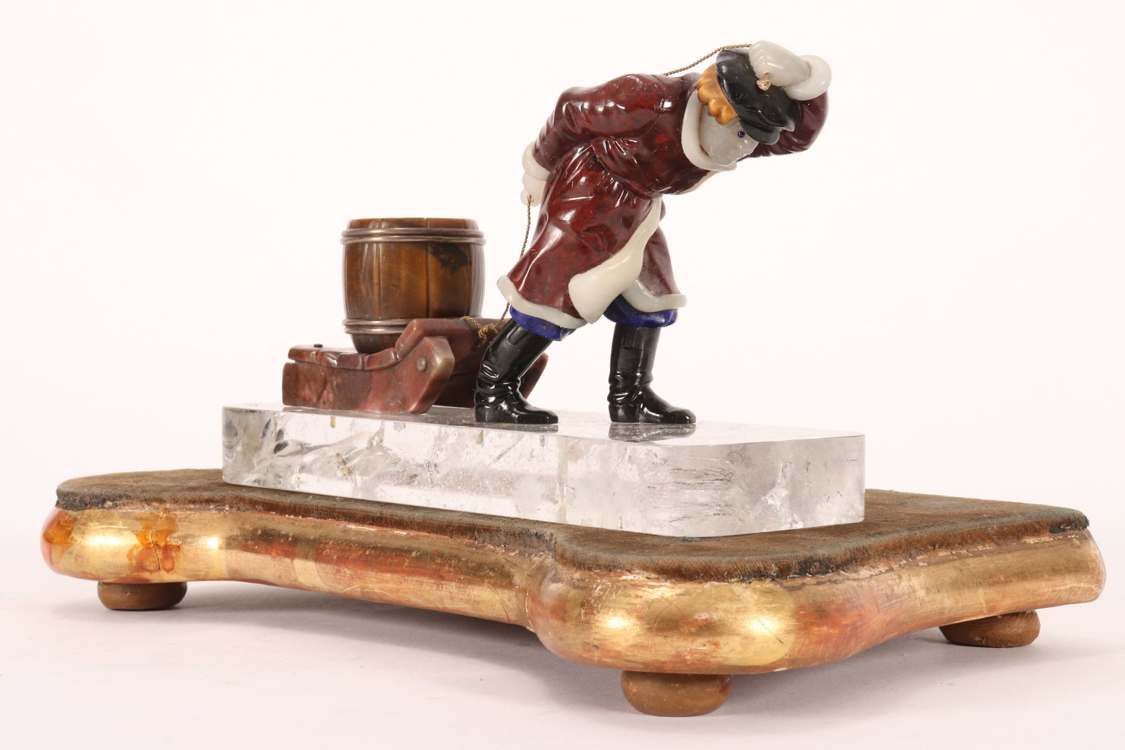 Russian Carved Stone Figure Group,: depicting a man in a winter coat and hat pulling a strapped barrel resting upon a sled, in tones of maroon, white, orange and brown, the whole resting upon a rock crystal base, with silver marked Fabe