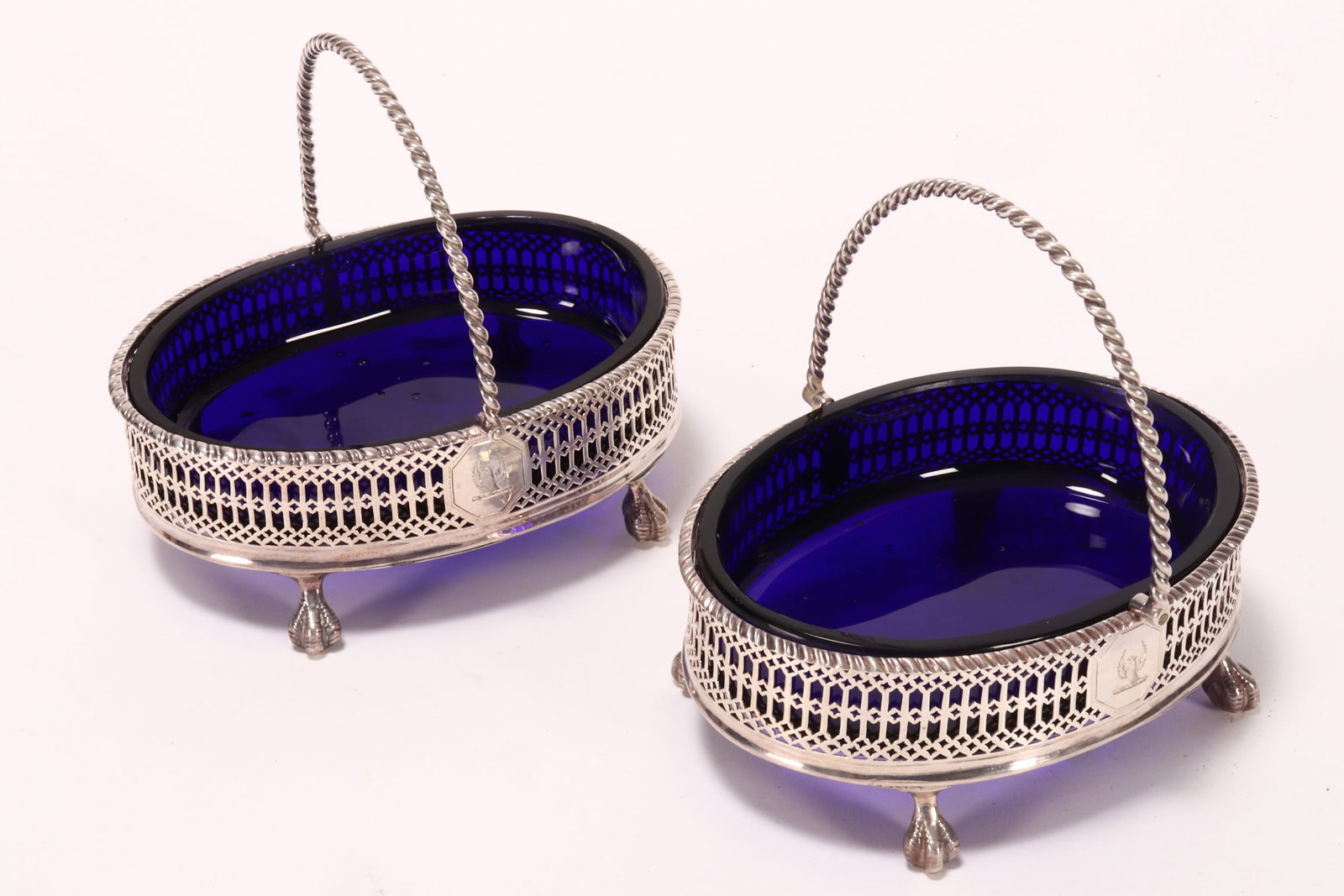 Fine Pair of George III Sterling Silver Footed Sweets Meats Dishes,: hallmarked London, 1774, T (makers mark rubbed), of oval form, with twist loop handle, the body finely pierced with cartouche crest, raised on ball and claw feet, set with cobalt glass liners, total w