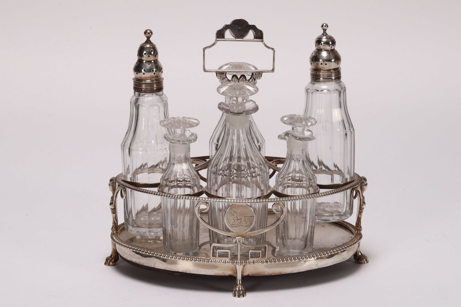 George III Sterling Silver and Crystal Cruet Set,: hallmarked London, 1806, makers mark rubbed, of elliptical form, set with tall central handle, the pierced frame decorated with two reserves containing engraved crests and stylised Egyptian busts, acc