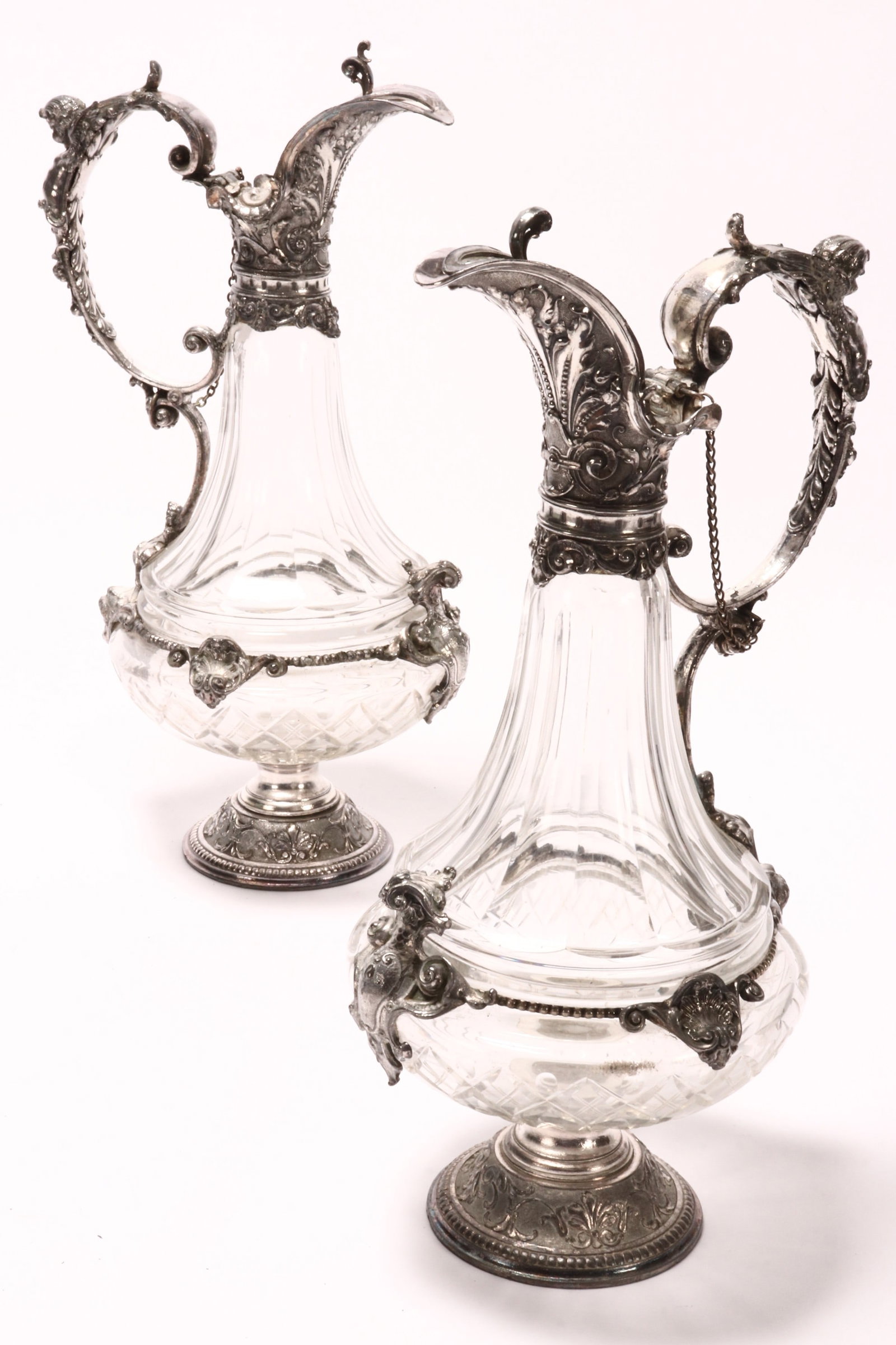 Pair of Silver Plate and Cut Glass Claret Jugs,: with hinged cover and elongated spout, decorated densely with scrolled acanthus, set with figural c-scroll putti handle, above a faceted pear form body, accented with shell, acanthus and bead border, 