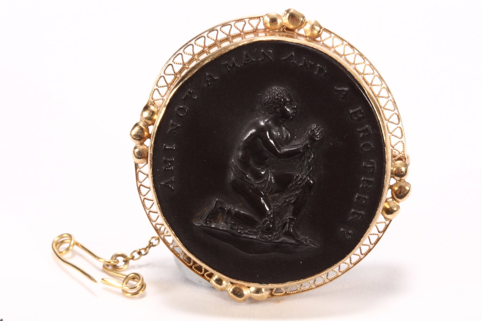 Wedgwood Black Basalt Abolitionist Brooch,: the cameo medallion reading 'Am I Not a Man and a Brother?', impressed Wedgwood//Made in England//58//EC verso, in 14ct gold stamped pierced setting, with gold safety chain and pin, 3.5cm x 3.5cm