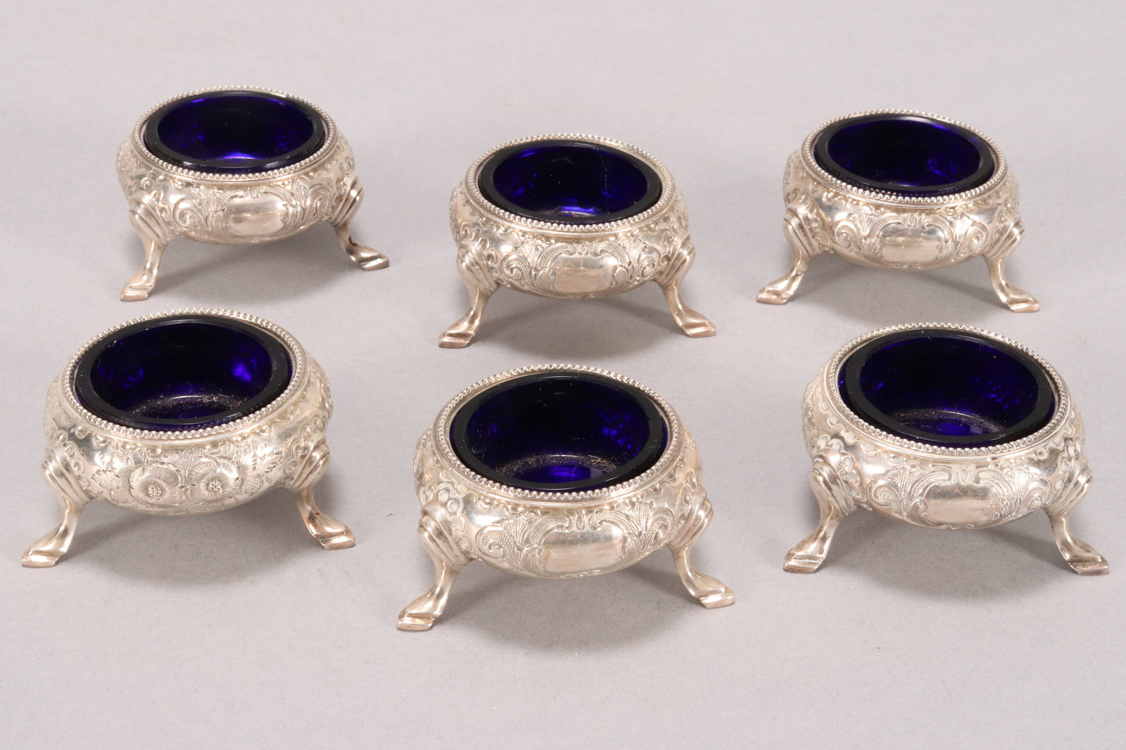 Set of Six Victorian Sterling Silver Salts,: hallmarked Sheffield, 1863, Martin and Hall, with fine beaded rims, of squat baluster form, embossed with floral motifs, raised on three hoof feet, with gilt interiors and blue glass liners, total wei