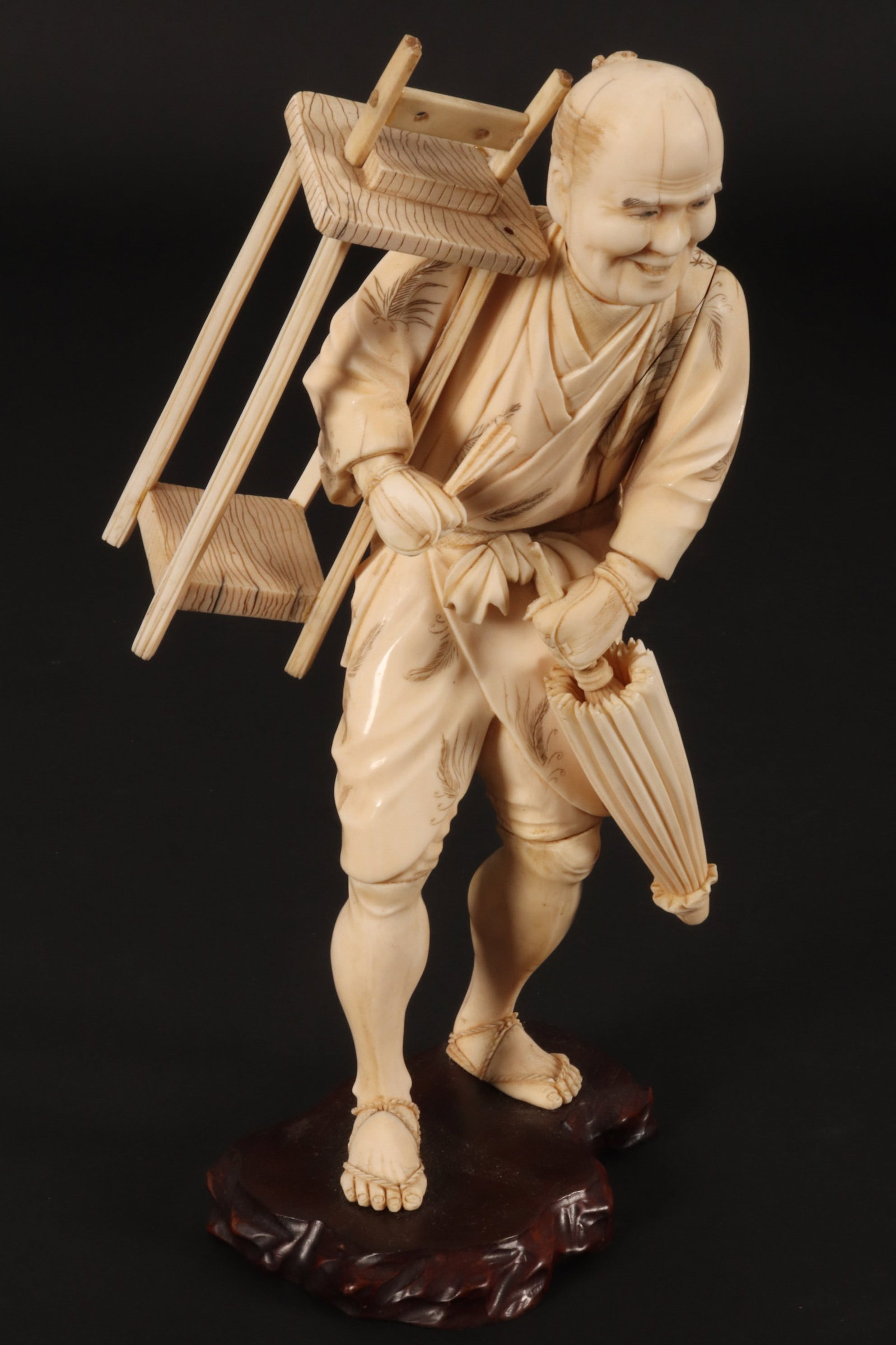 Large Japanese Meiji Period Figure,: depicting a man carrying a work table on his shoulder and holding a parasol, raised on a wooden base, height 27cm Please note this item can only be shipped WITHIN Australia.