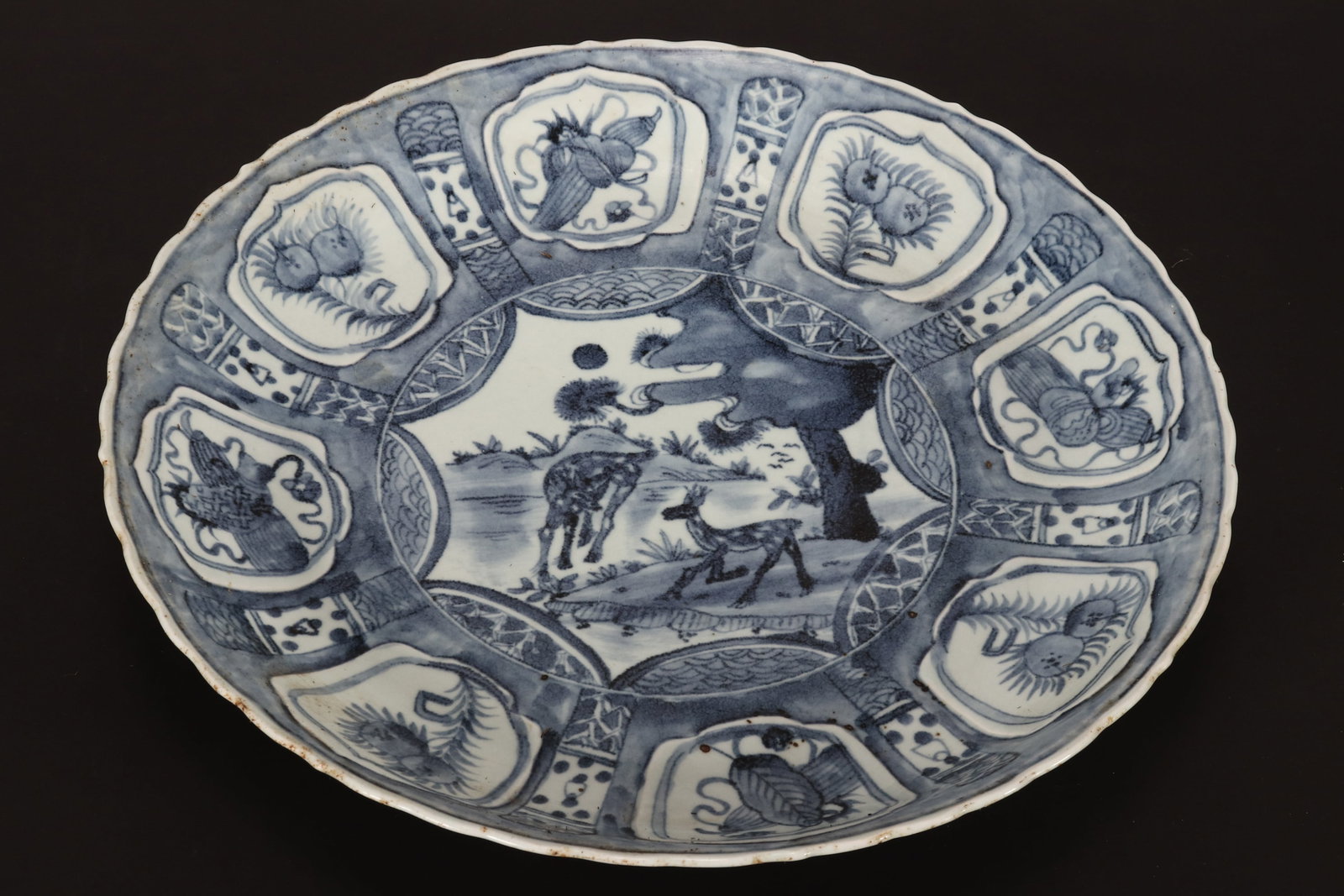 Chinese Late Ming Dynasty Kraak Charger,: in blue and white, with a serpentine edge, decorated with a central image of deer, surrounded by panels of fruit and objects, raised on a circular foot, diameter 39cm
