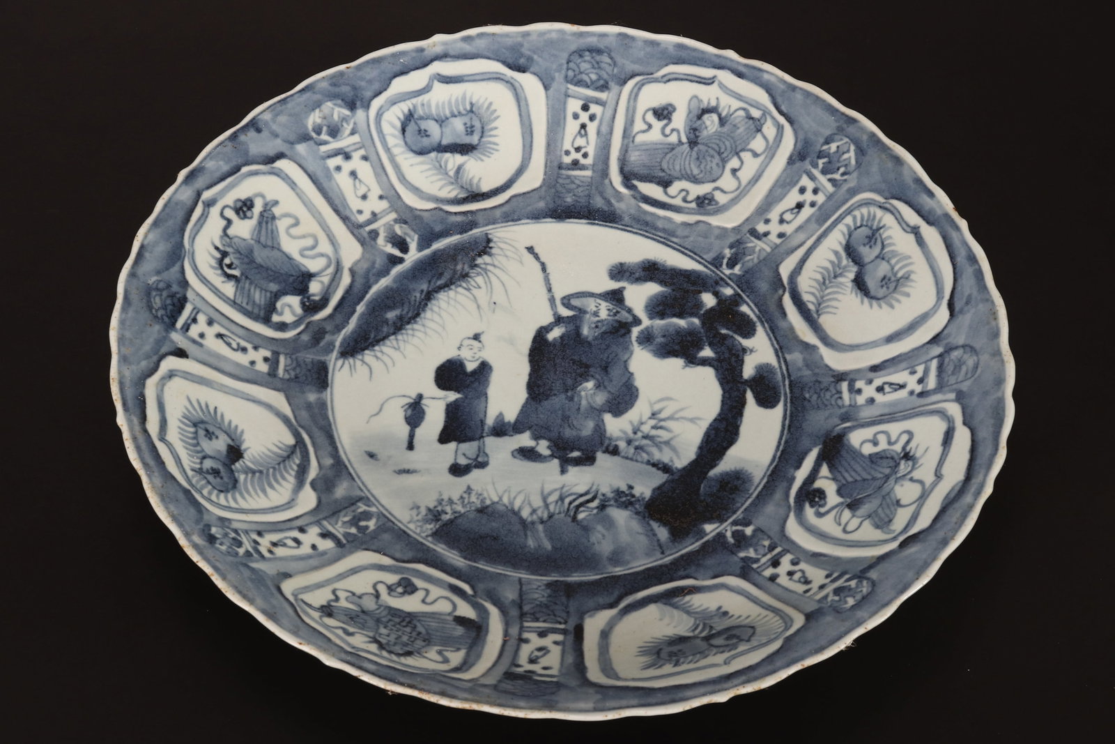 Chinese Late Ming Dynasty Kraak Charger,: in blue and white porcelain, with serpentine edge and central roundel of figures in landscape, surrounded by panels displaying fruit, raised on a circular foot, diameter 38cm