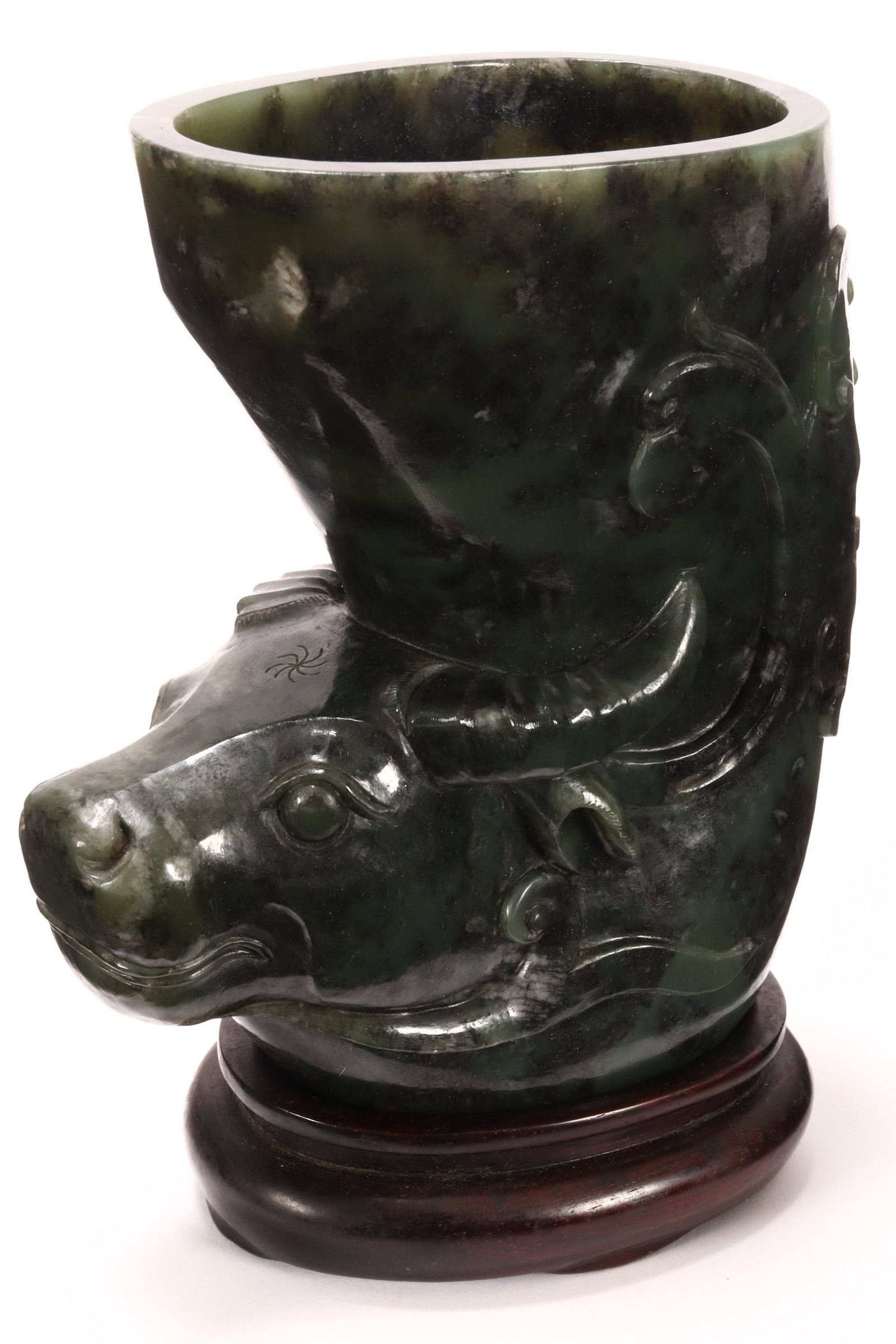 Chinese Green Jade Libation Cup on Stand,: of shaped oval form, decorated with water buffalo mask to body, below scrolls and foliage, in deep green and black tones, raised on a carved Chinese wooden stand, height 13.5cm and 15.5cm (including s
