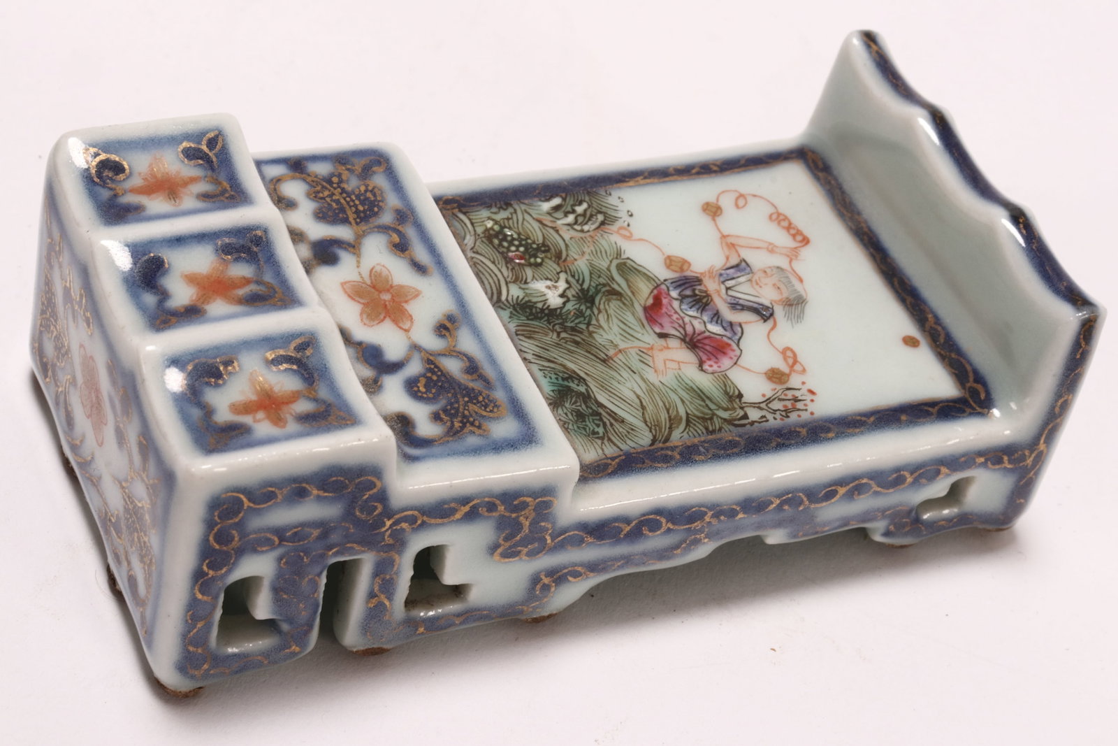 Chinese Porcelain Brush Rest,: of rectangular form, decorated with a panel depicting a seated figure holding rope tied with coins, surrounded by flowers and foliage, in tones of blue, green, orange and pink on a white ground, accen