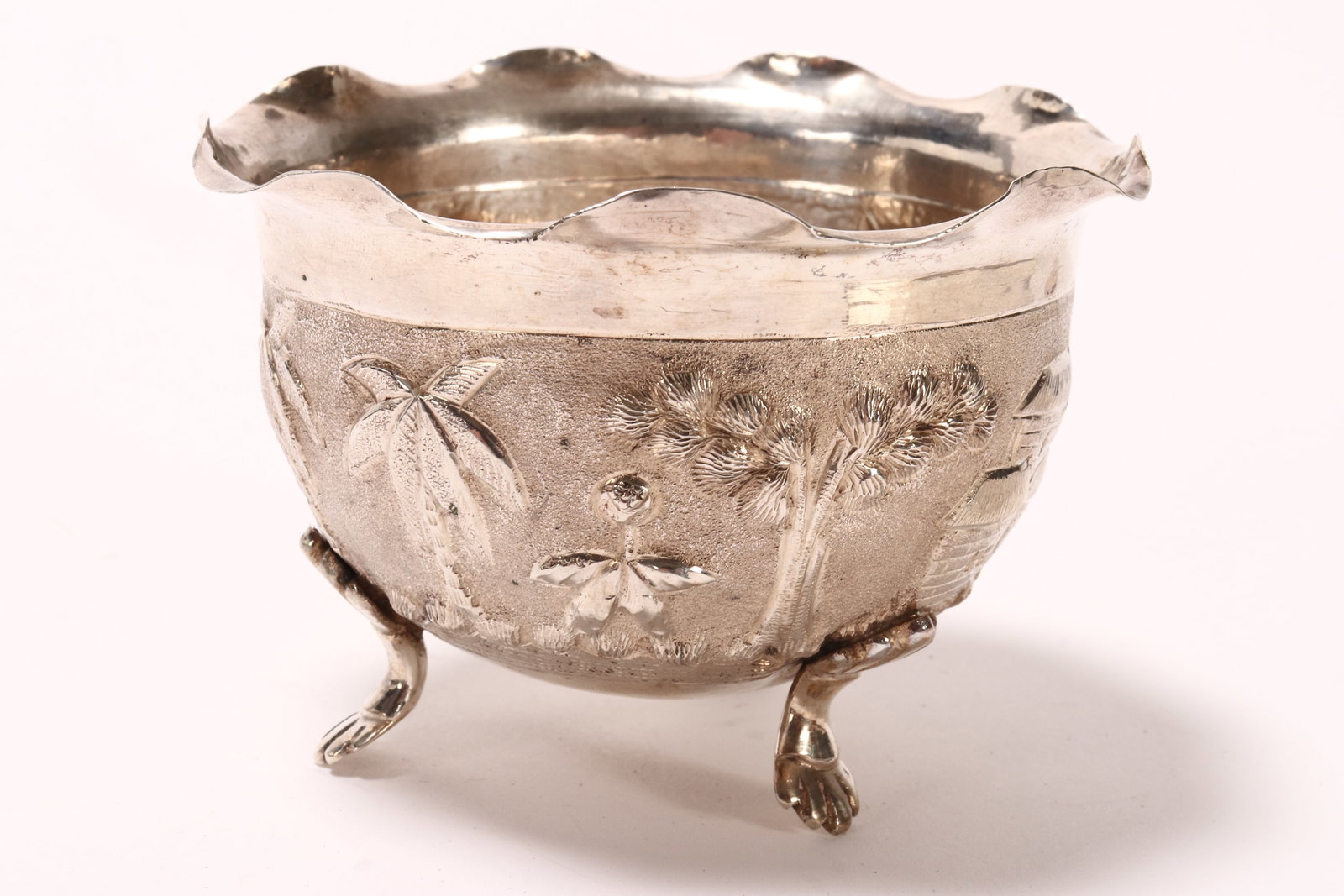 Burmese Silver Footed Bowl,: unmarked (acid tested silver), of deep form with shaped rim, the body decorated with embossed landscape with huts and trees, the whole raised on three shell form feet, total weight 63g, height 6cm