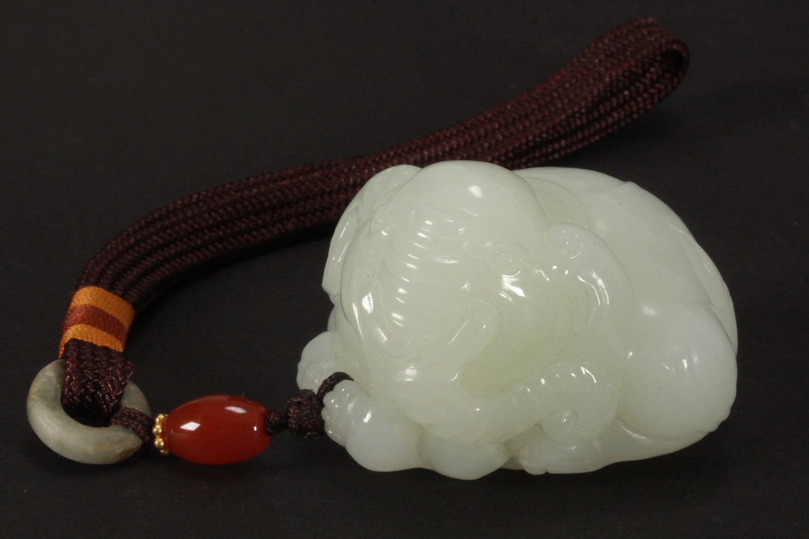 Chinese Carved Pale Green Jade Elephant,: depicted seated on all fours with a coiled tail and trunk, set on cord with red bead and jade disc, height 3.5cm length 5cm (elephant)