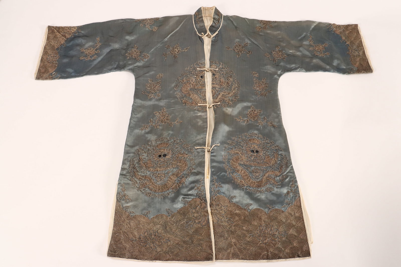 Chinese Embroidered Silk Surcoat,: the whole decorated with gold-work embroidered dragons in flight, amongst foliate sprays, above turbulent waves to the lower hem and sleeves, in silvered tones on a sky blue ground, set with white met