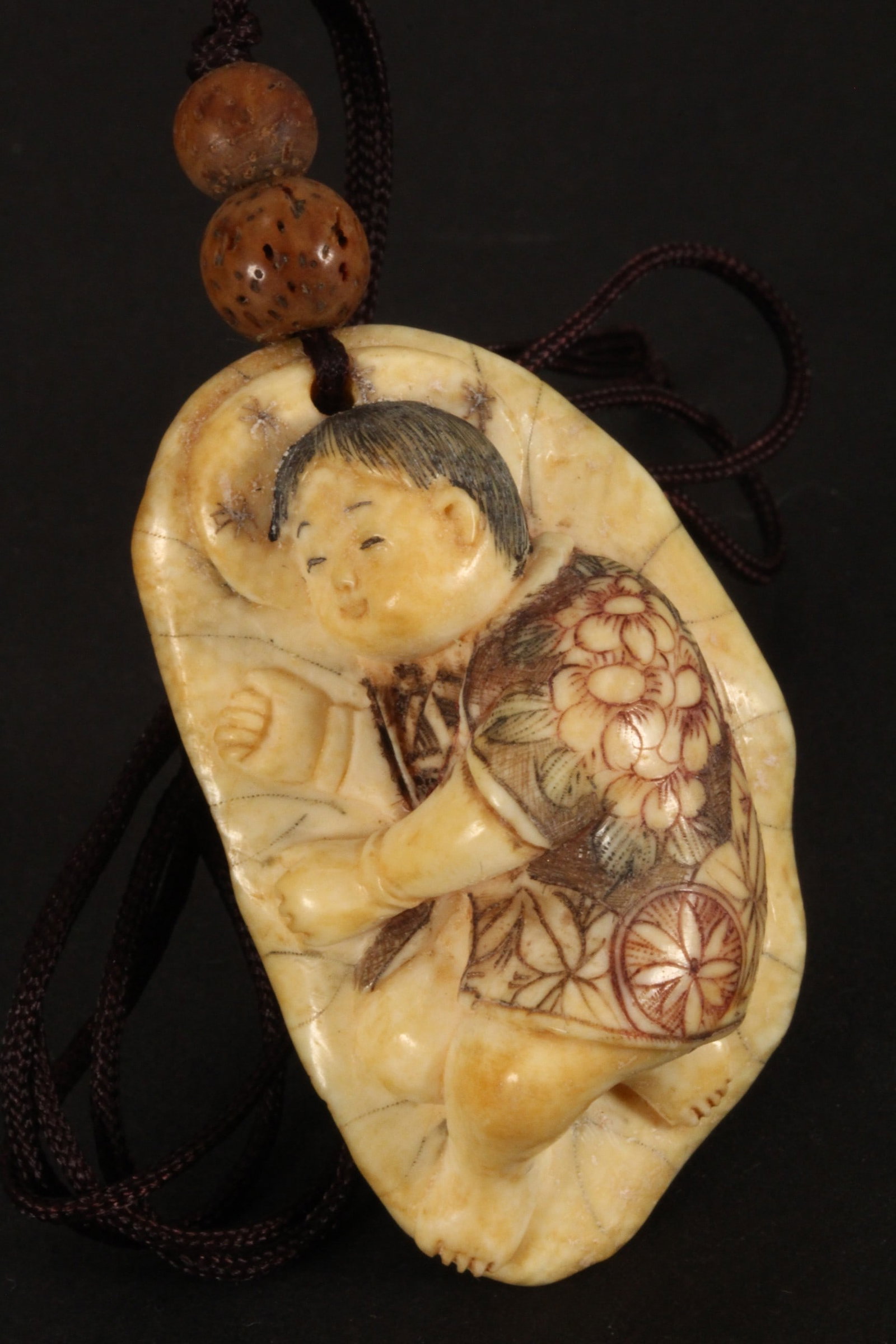 Japanese Carved Pendant,: in the form of a child sleeping on a lotus leaf, with incised and stained detail to the figure and clothing, suspended on a looped cord, incised signature to base, length 4.5cm. Please note this item 