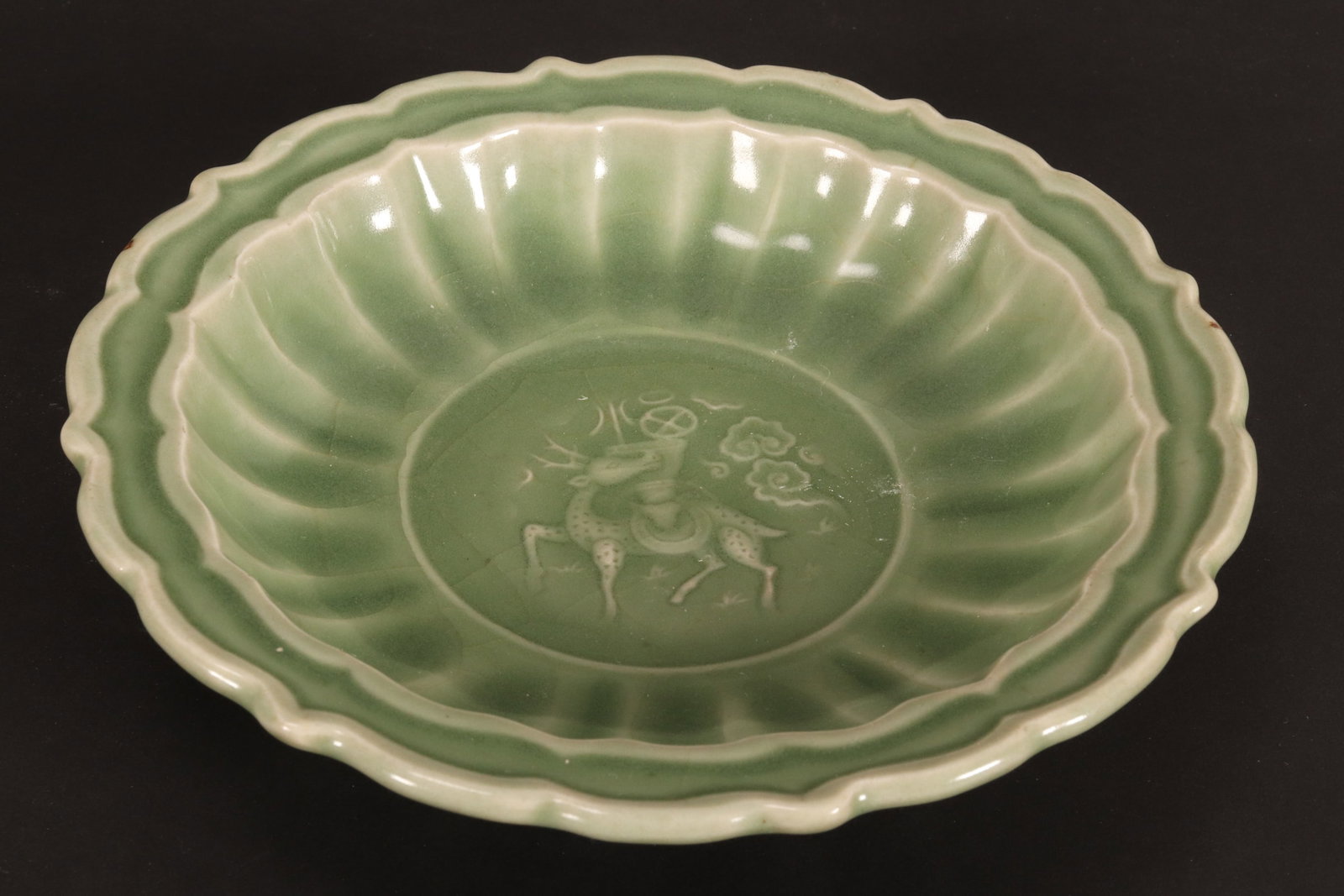 Chinese Celadon Dish,: of scalloped form, decorated with central roundel of a deer with stylised clouds and precious items, standing on a raised circular foot, diameter 19cm