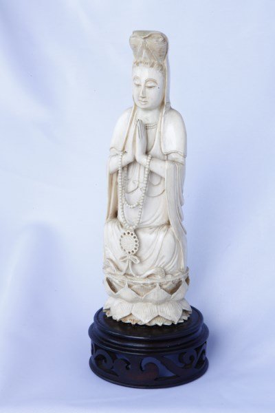 Good Chinese Carved Ivory Kwan Yin,: depicted with top knot and covering veil, hands placed together, with necklace hanging from hands, seated on double lotus, on carved wood stand, h 19cm, 23cm (with stand) Provenance: This item is one 