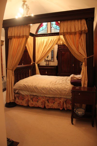 Late 19th Century Gothic Revival Four Poster Bed,: king size, with a stepped pediment, canopy and side curtains in a butter yellow brocade with tassel finish, the bed head and foot carved and pierced with a repeating ogee motif, the whole supported on