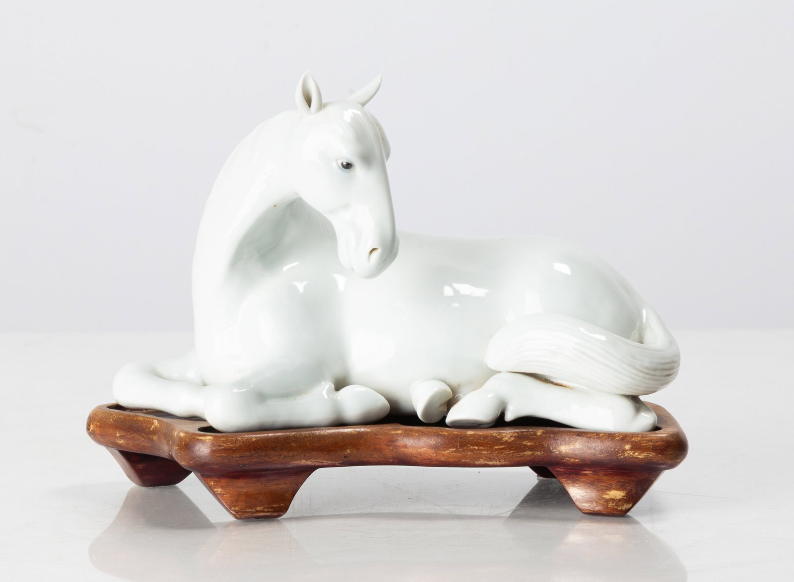 Chinese Porcelain Blanc de Chine Model of a Recumbent Horse: Chinese Porcelain Blanc de Chine Model of a Recumbent Horse China, Qing dynasty. Blanc de chine porcelain, on a fitted carved wood stand. The underside with a small impressed square seal and an inked