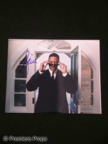Will Smith Signed Photo: This is a glossy, color autographed photo of actor Will Smith. The photo features Will Smith in black suit & tie, removing his trademark 'Men In Black' sunglasses, and is signed in blue felt ink. The