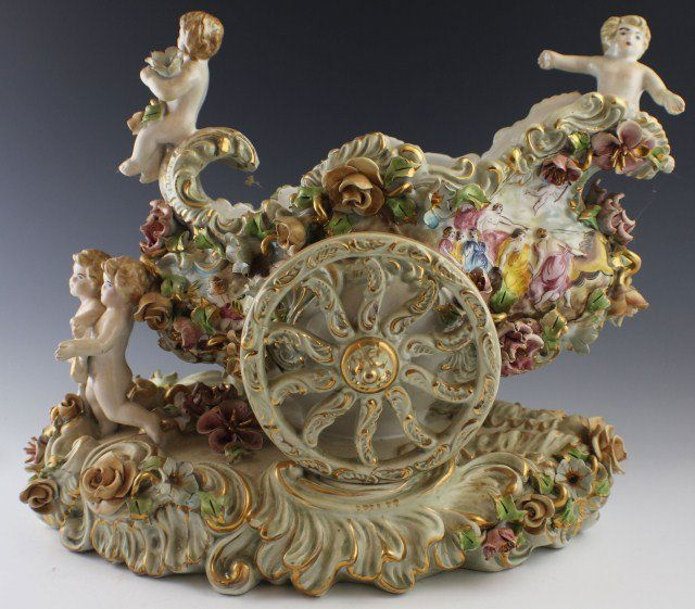 CAPODIMONTE PORCELAIN WHEELED VASE: CAPODIMONTE PORCELAIN WHEELED VASE Large Capodimonte center table vase depicted having a chariot wheel on each side. Two cherub figures are seated on top of the vase and two are leading it. Throughout