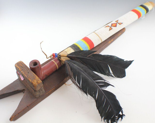 BEADED CEREMONIAL CATLINITE PIPE: BEADED CEREMONIAL CATLINITE PIPE Ceremonial beaded pipe mounted on a wooden wall display stand. Pipe is decorated by black, blue, yellow and white beading around the shaft, and is inlaid into a catlin