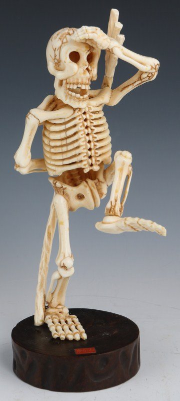 CARVED IVORY STANDING SKELETON SIGNED: CARVED IVORY STANDING SKELETON SIGNED Well executed ivory skeleton in mid stride high step salute, caring an ivory pitchfork. Small red plaque on 1" wooden base signed in Chinese. This item was origin