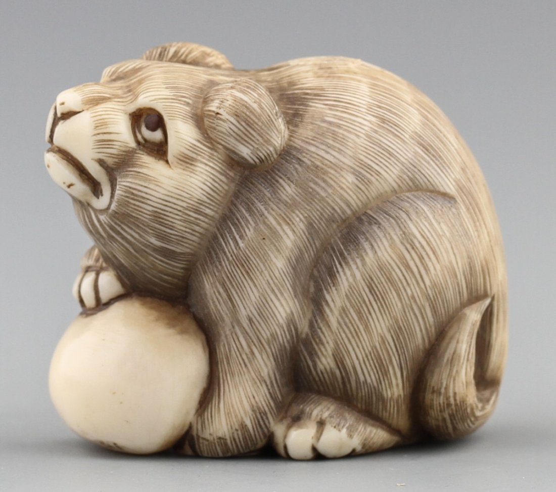 JAPANESE CARVED IVORY NETSUKE DOG BY BAISHO: JAPANESE CARVED IVORY NETSUKE DOG BY BAISHO Early 20th century Japanese ivory carved netsuke of dog with ball. Monochromed details, signed Baisho. Size: 1.25" Weight: 29g