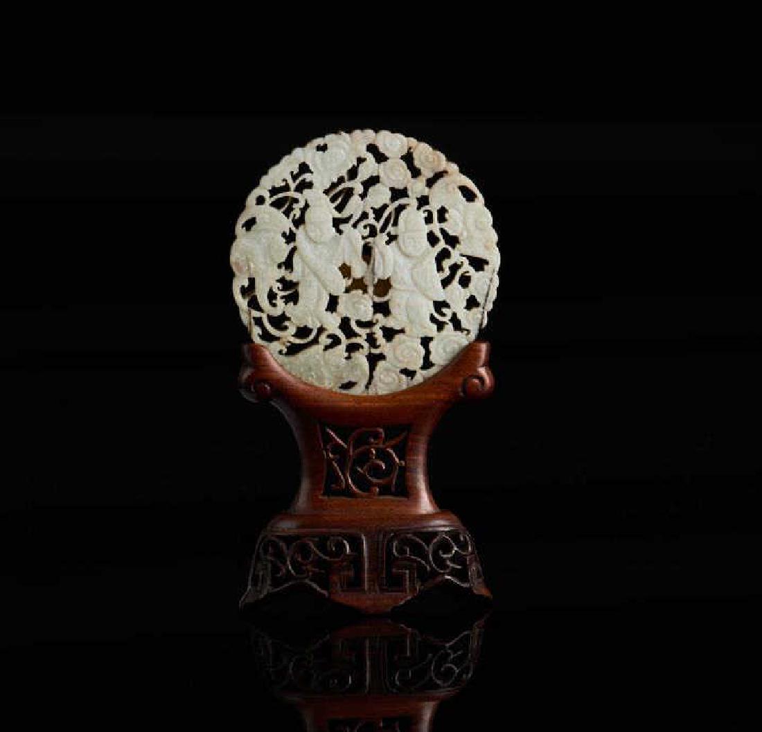 CHINESE JADE CARVED CIRCULAR PLAQUE: Overall in opaque white chicken bone tone, with brown skimming to one side. Carved with a man and a woman both accompanied by bats, clouds, and lotus scrolls. With fitted wood stand. Qing dynasty. Dia