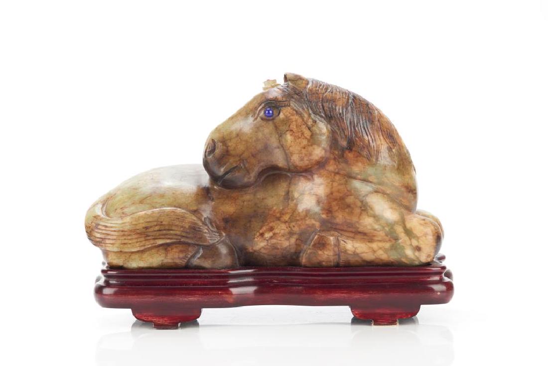 LARGE CELADON JADE CARVED RECLINING HORSE: Overall in celadon tone with russet brown inclusions. Carved as a reclining horse with blue inlaid eyes. With fitted wood stand. Four character seal mark to base. L. 30cm (12 in.)