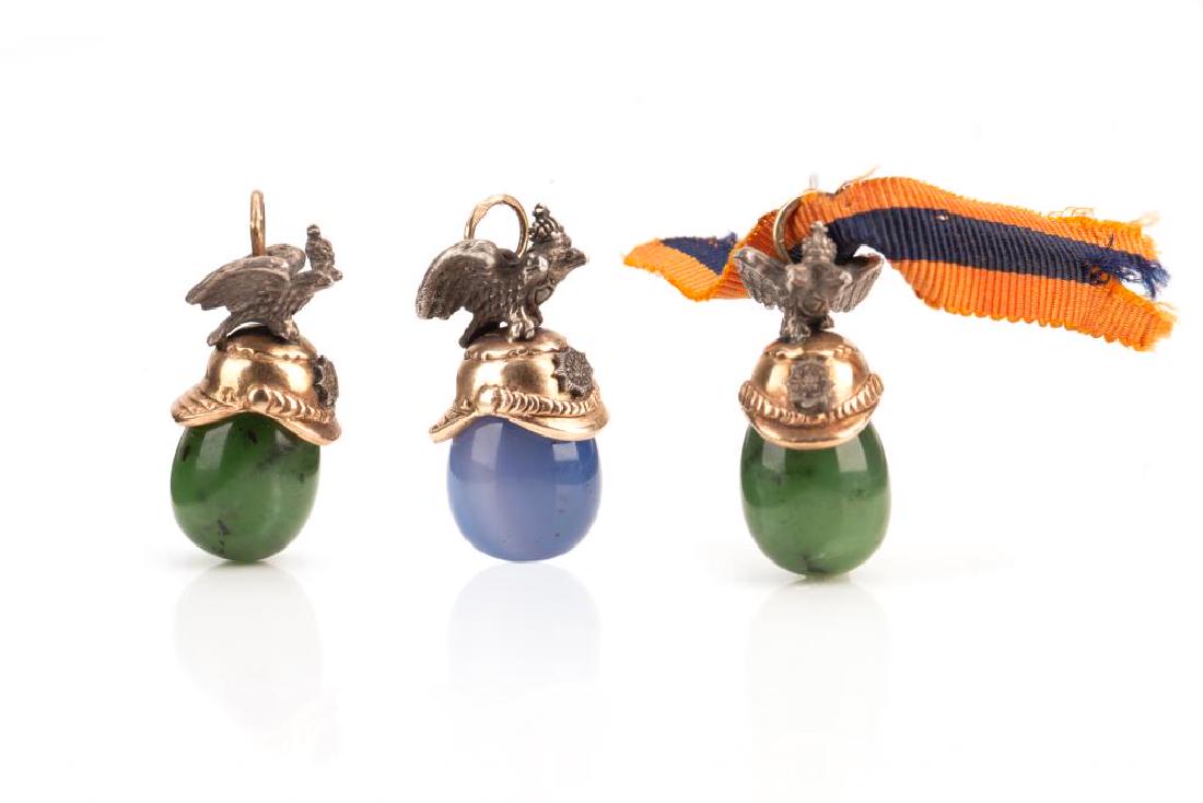 Three Russian silver and hardstone egg pendants: Each in the form of a cavalier novelty gold helmet, with a Habsburg eagle atop. Two with nephrite green jade and one in blue agate. The loops stamped 56.