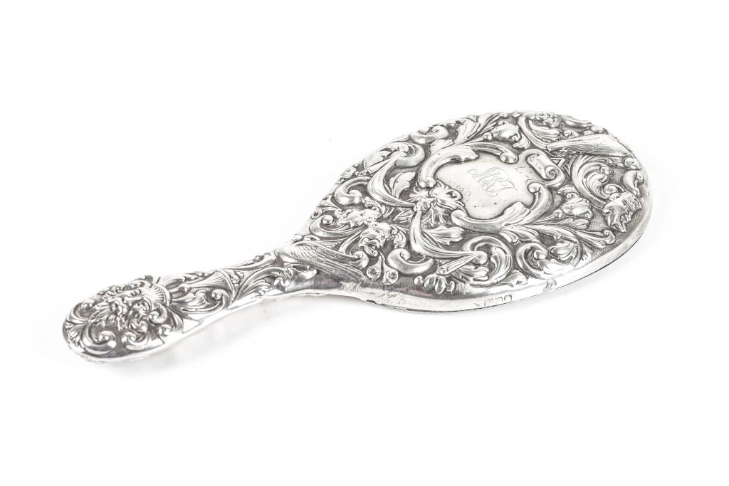 ENGLISH SILVER REPOUSSE HAND MIRROR: A late Victorian handheld mirror mounted in sterling silver with elaborate detailing in high relief of foliage, fruits, and faces surrounding a central monogrammed cartouche on back. Mirror in oval fo