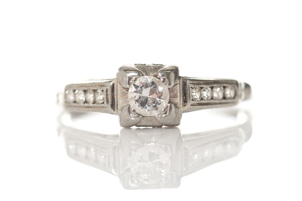 WHITE GOLD AND DIAMOND RING, 3g: Late Edwardian white gold ring with a central prong set round diamond. Each shoulder flank has four round cut channel set diamonds. Stamped 18k. Size 8. With case. Weight: 3g