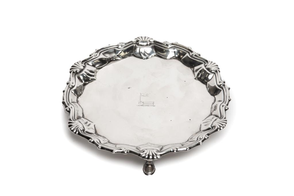 MID 18TH C SILVER SALVER, 218g: A Provincial or colonial salver on three hoof feet with cast and applied shell border bearing pseudo-hallmarks. Typical 1740s design. Engraved in the centre with a mailed arm holding a poignard tipped