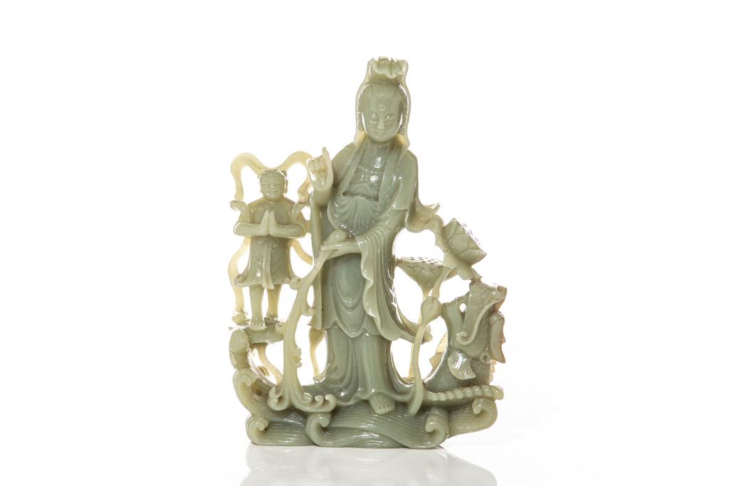 LARGE JADE CARVED GUANYIN AND BOY GROUP: Overall in celadon tone carved with a standing Guanyin figure accompanied by a boy attendant both riding a wave with dragon. H. 26cm (10 1/4 in.) Condition report available upon request.