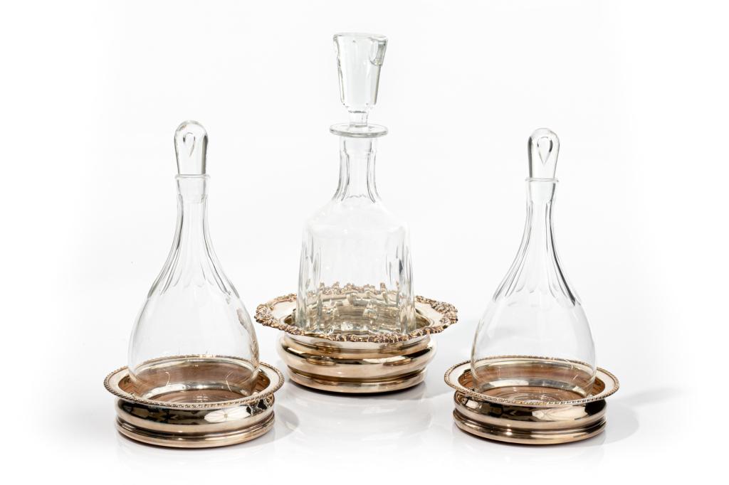 THREE SILVERPLATE COASTERS WITH GLASS DECANTERS: Two smaller coasters have gadrooned style border, the larger with a foliate border. Each with wood base. Decanters each have stoppers.H. of tallest decanter 29 cm (11 1/2 in) Dia. of largest coaster 1