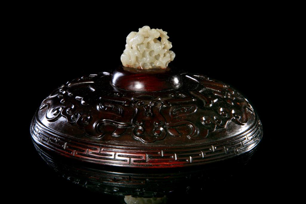 CHINESE WOOD COVER WITH JADE FINIAL: Finial of a finely carved jade official's hat finial, Yuan dynasty. Cover carved with relief taotie mask motifs. 20th C. Cover dia. 19.5cm (7 3/4 in.) H. of jade: 3.5cm (1 3/8 in.)