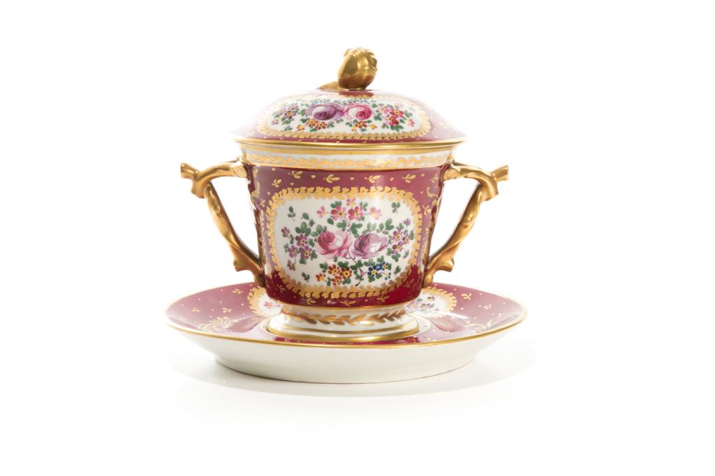 LIMOGES PORCELAIN LIDDED POT ON STAND: French porcelain, berry red ground with gilded accents and hand painted floral panels. Footed jar with double gilded handles and rosebud finial to lid, with under plate stand. Makers marks to the unde