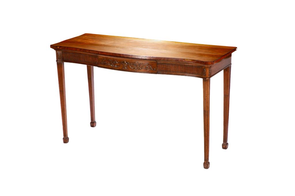 GEORGE III ADAM FORM MAHOGANY SERVER TABLE: Single drawer, serpentine surface with carved neoclassical motifs in relief. With fluted tapered square legs. English, second half 18th C. H. 35" W. 58" D. 24" Provenance: The Estate of the Late Senat