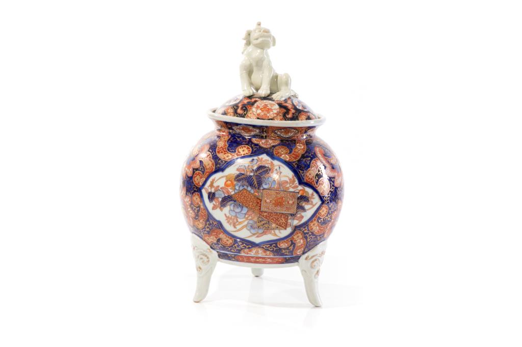 JAPANESE IMARI PORCELAIN TRIPOD KORO CENSER: Cover with a sculpted shishi lion form finial. Body painted in Imari palette with decorative floral motifs. Tripod foot with mask tops. Meiji period. H. 38cm (15 in.) Provenance: From Chelster Hall Oa